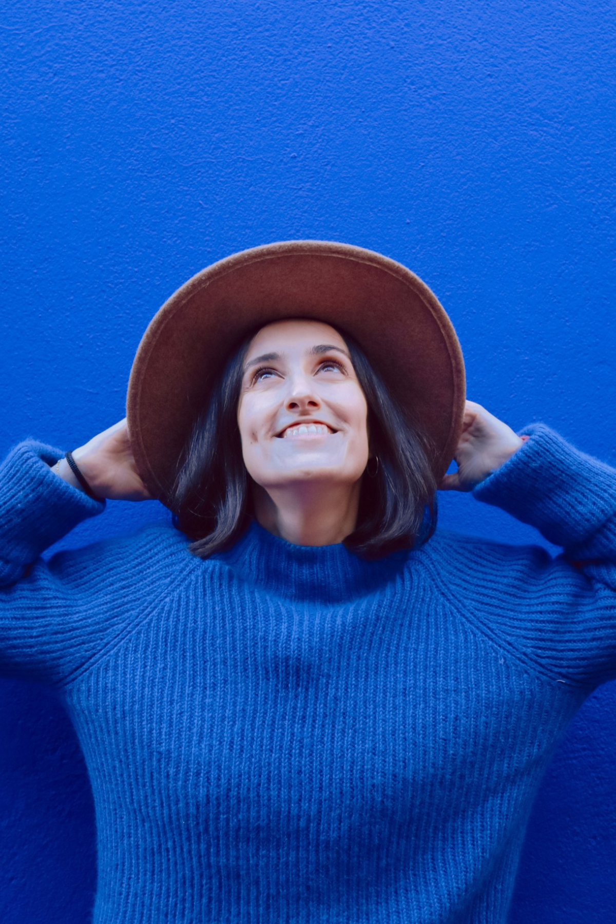 Mariana smiling in a brown hat and blue sweater against a bright blue wall